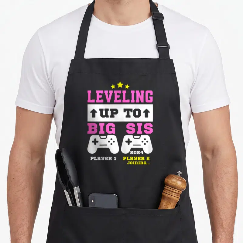 Tablier de cuisine noir avec message Leveling Up to Big Sis 2024 et deux manettes de jeu vidéo blanches.