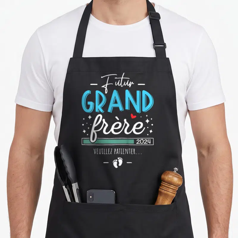 Tablier de cuisine noir avec l'inscription Futur Grand frère 2024 Veuillez patienter et des empreintes de pieds en bas du motif.