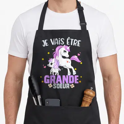Tablier de cuisine noir avec illustration de deux licornes roses et le texte Je vais être grande soeur entouré d'étoiles.