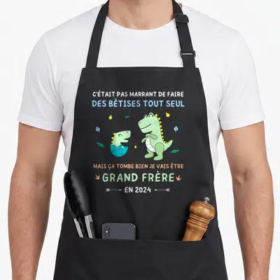 Tablier de cuisine noir avec un dinosaure vert et l'annonce Grand Frère en 2024.
