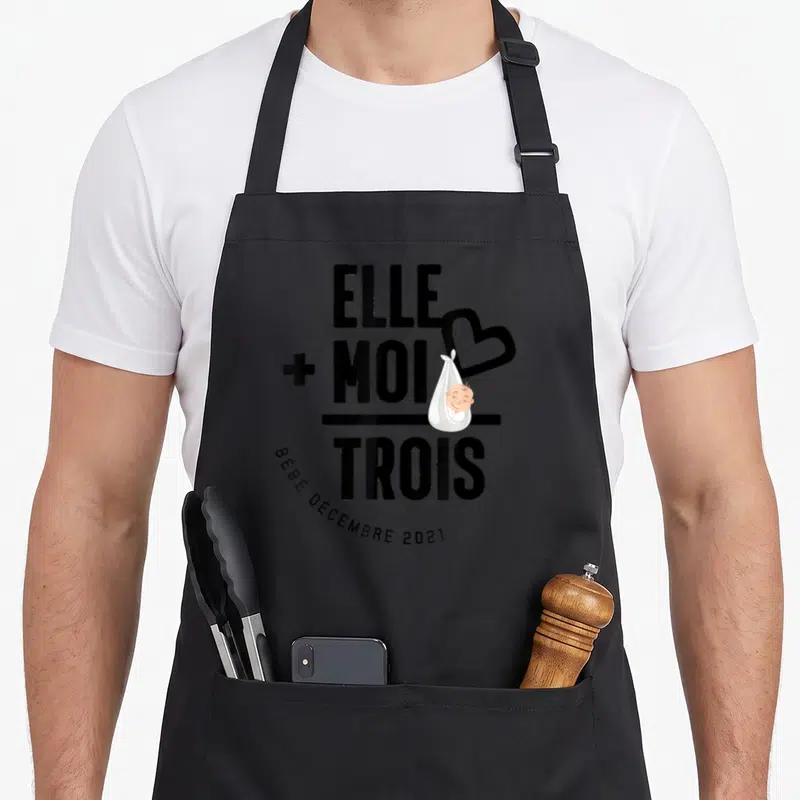 Tablier de cuisine noir avec le texte Elle plus Moi trois et une illustration de bébé dans un sac blanc.