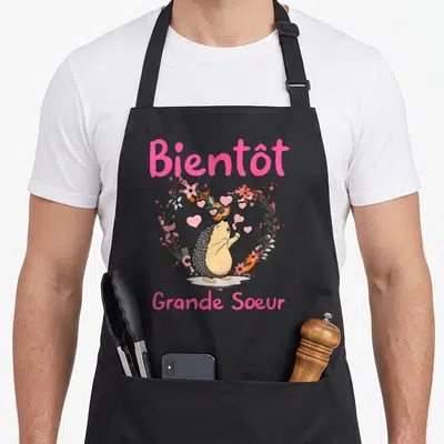 Tablier de cuisine noir avec un hérisson tenant une loupe sous un cœur floral et le texte Bientôt Grande Soeur en rose.