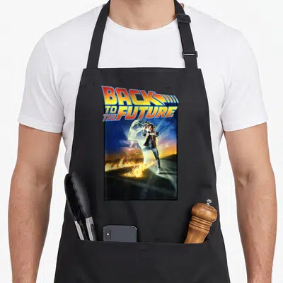 Tablier de cuisine noir avec l'affiche rétro de Back to the Future montrant Marty McFly et la voiture temporelle en feu.
