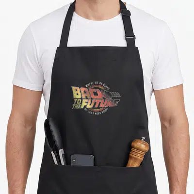 Tablier de cuisine noir avec logo rétro Back to the Future et texte jaune orange sur fond gris uni.