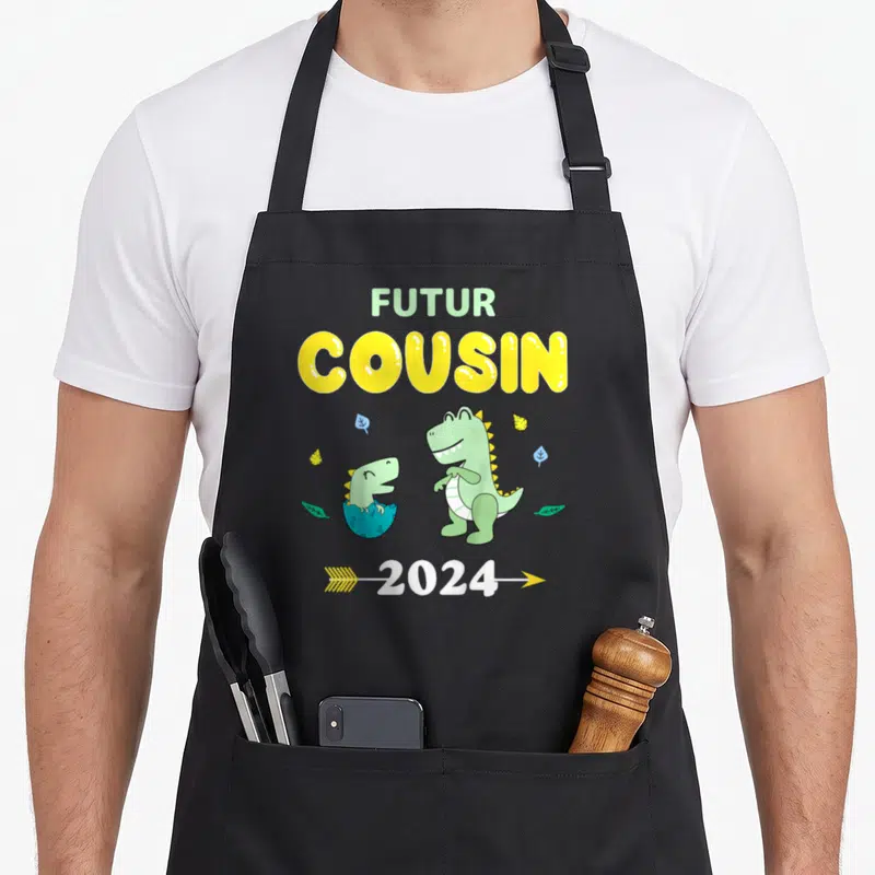 Tablier de cuisine noir avec l'inscription Futur Cousin 2024 et deux dinosaures verts souriants entourés de feuilles.