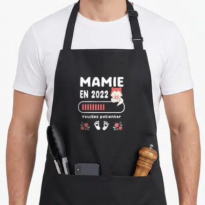 Tablier de cuisine noir avec inscription Mamie en 2022, illustration d'un bébé et barre de chargement rose.