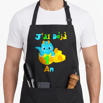 Tablier de cuisine noir avec un dinosaure bleu sortant d'une coquille jaune et le texte J'ai déjà 1 an en lettres colorées.