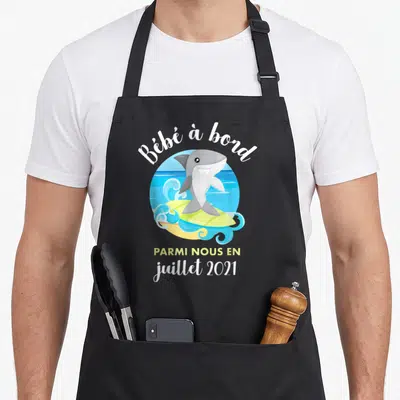 Tablier de cuisine noir avec un requin souriant sur une planche de surf et le texte Bébé à bord parmi nous en juillet 2021.