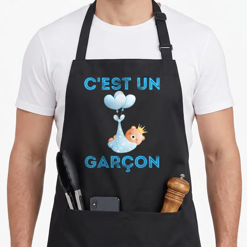 Tablier de cuisine noir avec illustration d'un ours en couronne dans un drap bleu attaché à des ballons et le texte C'est un Garçon.