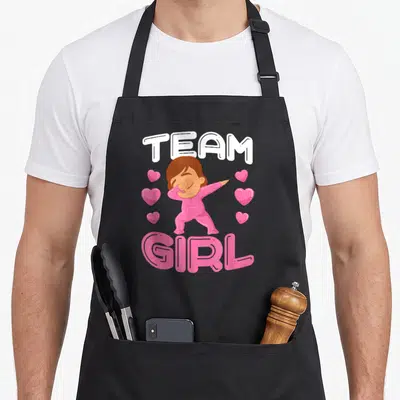 Tablier de cuisine noir avec un bébé en pink doing dab et les mots Team Girl entourés de cœurs roses brillants.
