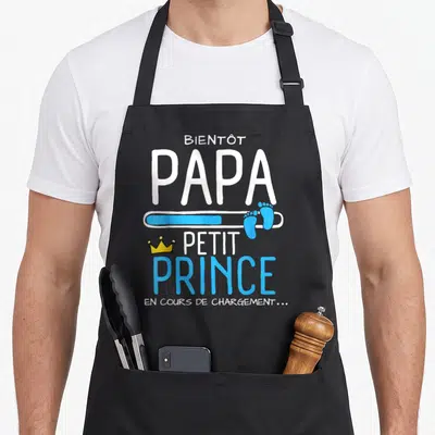 Tablier de cuisine noir avec l'inscription Bientôt Papa Petit Prince et une barre de chargement bleue ornée de petits pieds.