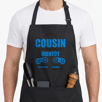 Tablier de cuisine noir avec texte bleu Prépare-toi cousin j'arrive et deux manettes de jeu vidéo.