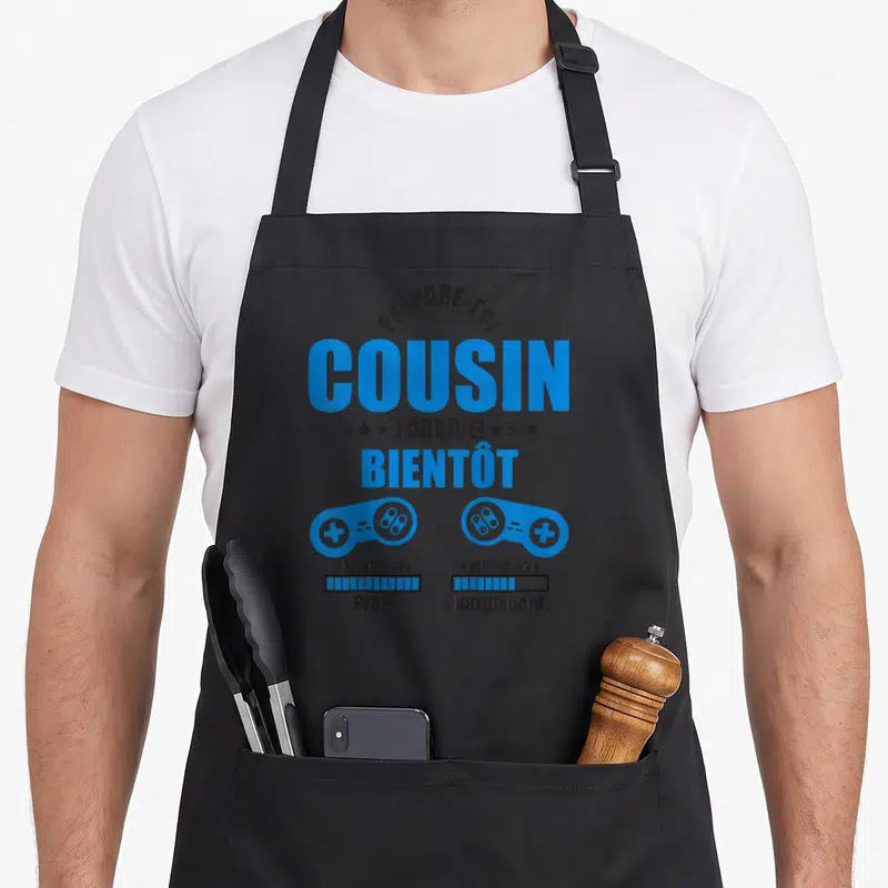 Tablier de cuisine noir avec texte bleu Prépare-toi cousin j'arrive et deux manettes de jeu vidéo.