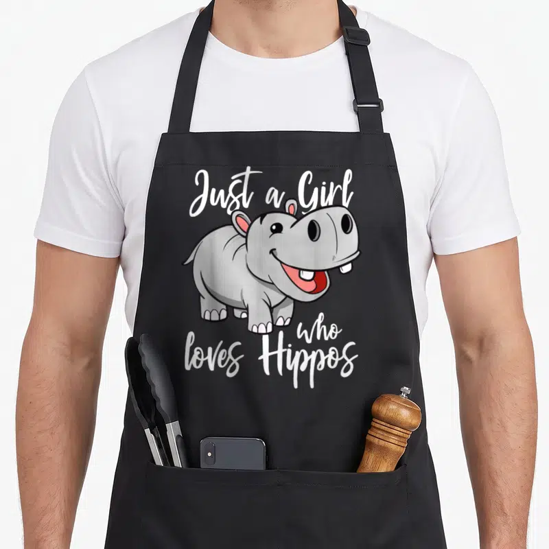 Tablier de cuisine noir avec un dessin de bébé hippopotame gris souriant et du texte blanc en anglais.