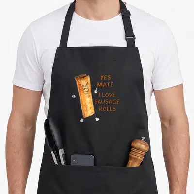 Tablier de cuisine noir avec un saucisson anthropomorphé souriant et du texte orange Yes Mate I Love Sausage Rolls.