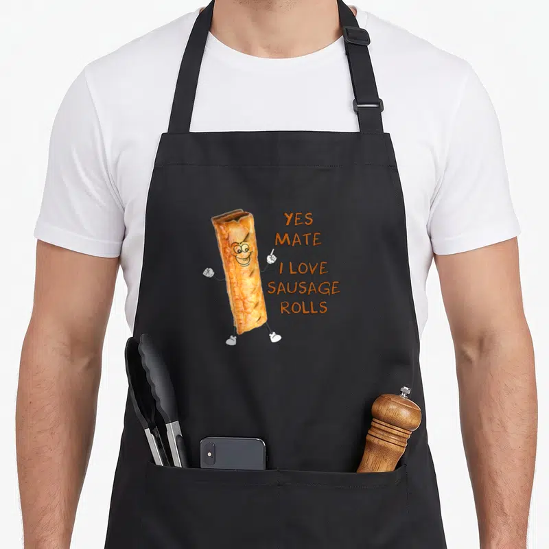 Tablier de cuisine noir avec un saucisson anthropomorphé souriant et du texte orange Yes Mate I Love Sausage Rolls.