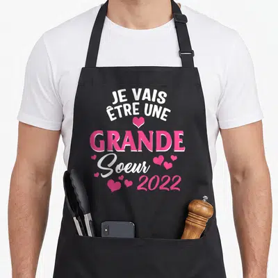 Tablier de cuisine noir avec l'annonce Je vais être une grande soeur 2022 en rose et blanc entouré de petits cœurs.