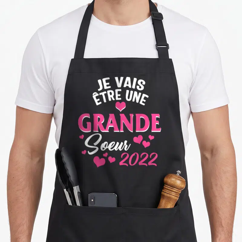 Tablier de cuisine noir avec l'annonce Je vais être une grande soeur 2022 en rose et blanc entouré de petits cœurs.