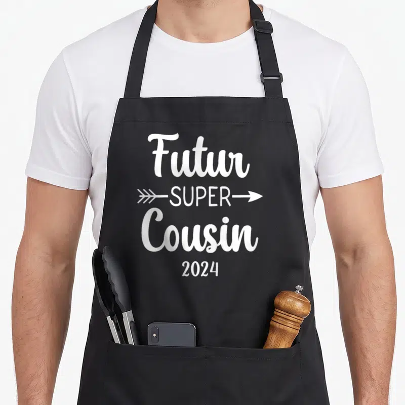 Tablier de cuisine noir avec inscription blanche Futur Super Cousin 2024 et flèches directionnelles.