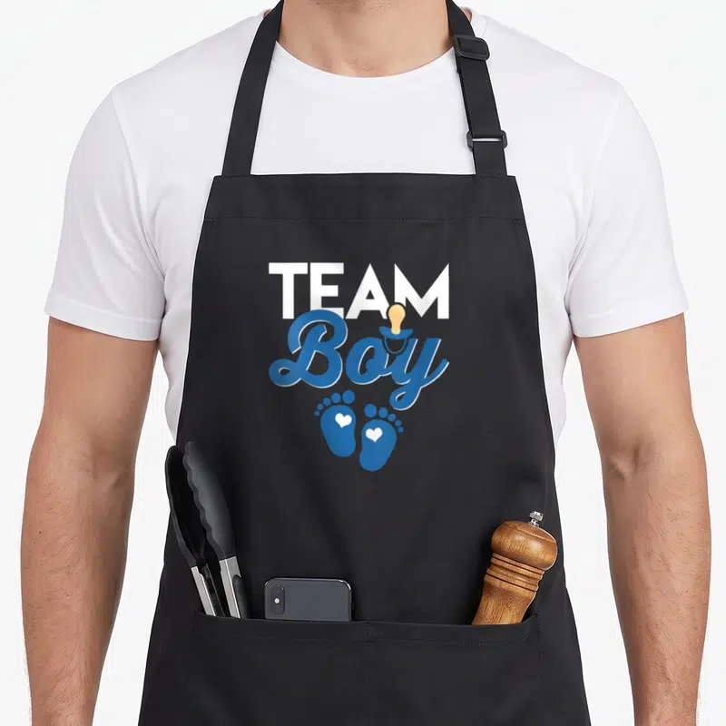 Tablier de cuisine noir avec l'inscription Team Boy, une sucette et deux empreintes de pieds bleues contenant des cœurs blancs.