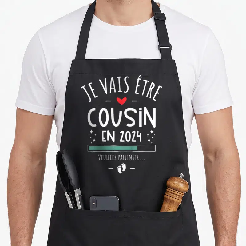 Tablier de cuisine noir avec texte blanc Je Vais Être Cousin En 2024, cœur rouge et petits pieds imprimés.