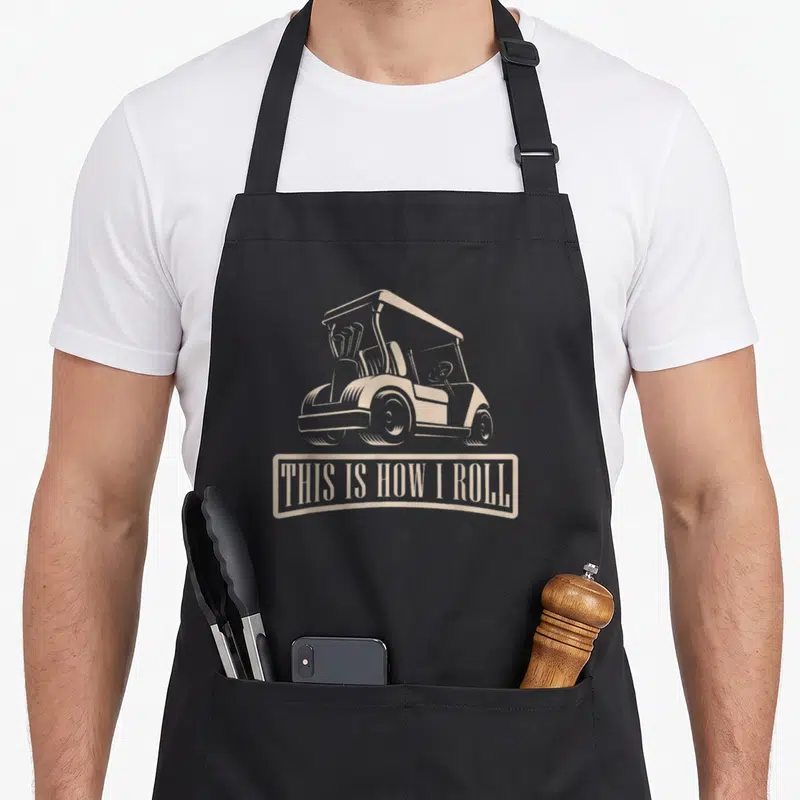 Tablier de cuisine noir avec illustration de caddie de golf beige et texte This Is How I Roll en bas.