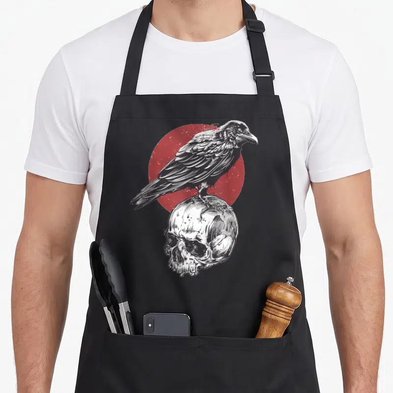 Tablier de cuisine noir avec illustration d'un corbeau posé sur un crâne devant un cercle rouge texturé.