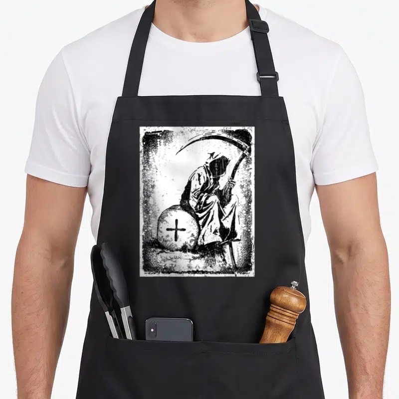 Tablier de cuisine noir avec une illustration gothique représentant une Faucheuse assise près d'une croix sur fond texturé.