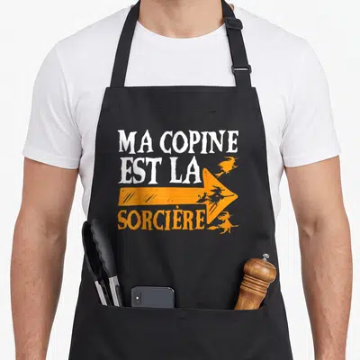 Tablier de cuisine noir avec texte blanc et flèche orange portant des silhouettes de sorcières volantes.