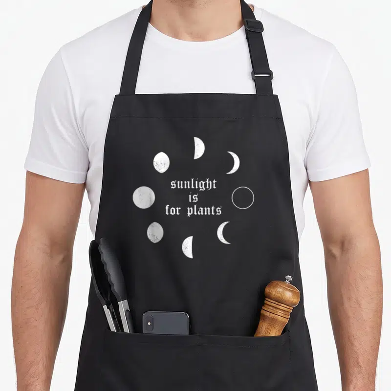 Tablier de cuisine noir avec texte sunlight is for plants et phases lunaires blanches en style grunge.