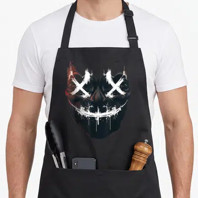 Tablier de cuisine noir avec masque effrayant aux yeux barrés et sourire blanchâtre peint au pochoir.