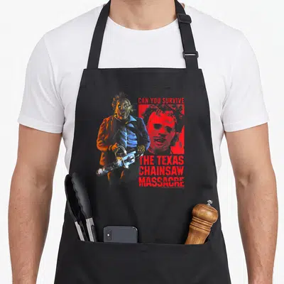 Tablier de cuisine noir avec illustration colorée de Leatherface tenant une tronçonneuse et un texte rouge en anglais.