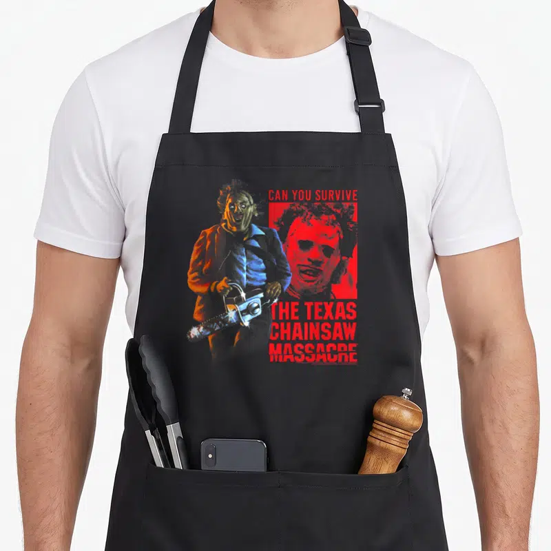 Tablier de cuisine noir avec illustration colorée de Leatherface tenant une tronçonneuse et un texte rouge en anglais.