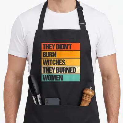 Tablier de cuisine noir avec inscription They Didn't Burn Witches They Burned Women sur fond de bandes horizontales orange et turquoise.