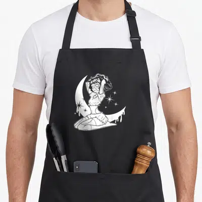 Tablier de cuisine noir avec une illustration blanche d'une poupée gothique assise sur un croissant de lune entouré d'étoiles scintillantes.