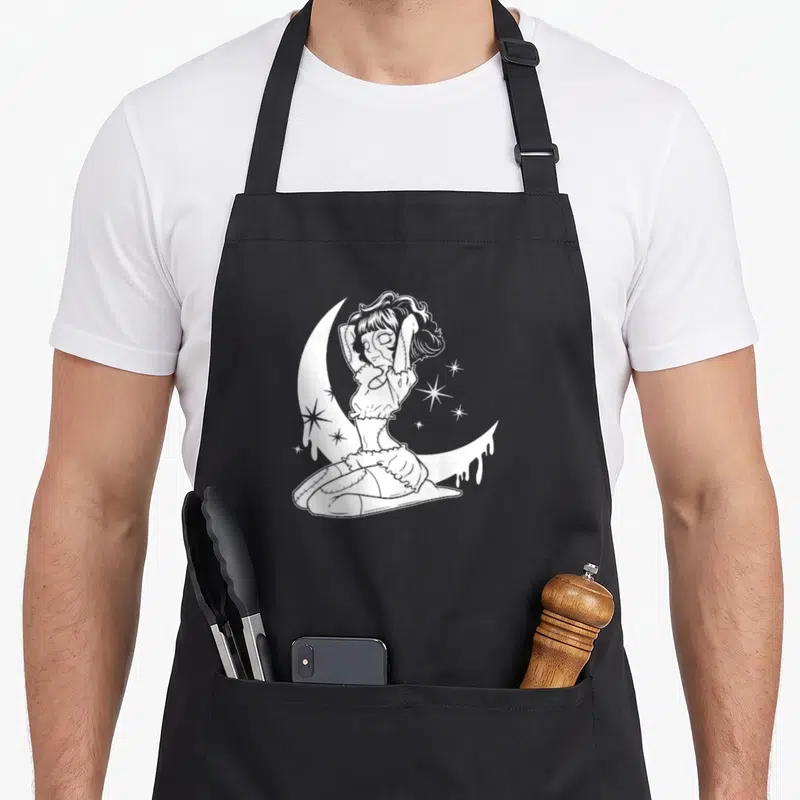 Tablier de cuisine noir avec une illustration blanche d'une poupée gothique assise sur un croissant de lune entouré d'étoiles scintillantes.