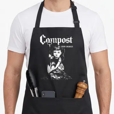 Tablier de cuisine noir avec illustration d'une fille tenant une pelle et le texte Compost Your Enemies en typographie gothique blanche.