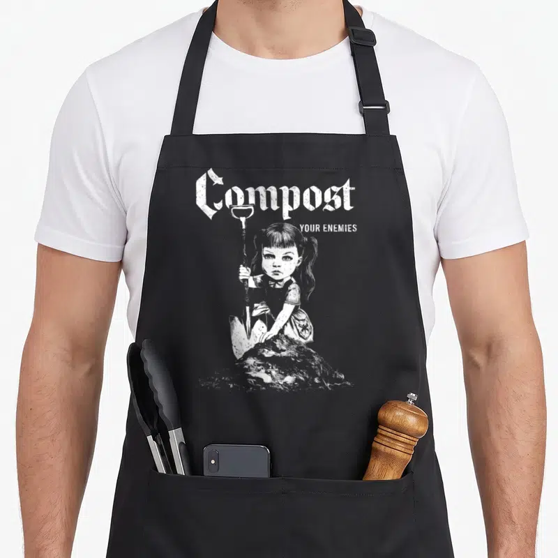 Tablier de cuisine noir avec illustration d'une fille tenant une pelle et le texte Compost Your Enemies en typographie gothique blanche.