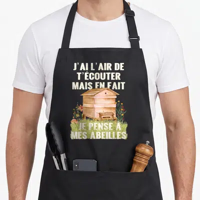 Tablier de cuisine noir avec une ruche en bois, des fleurs colorées et le texte J'ai l'air de t'écouter mais en fait je pense à mes abeilles.