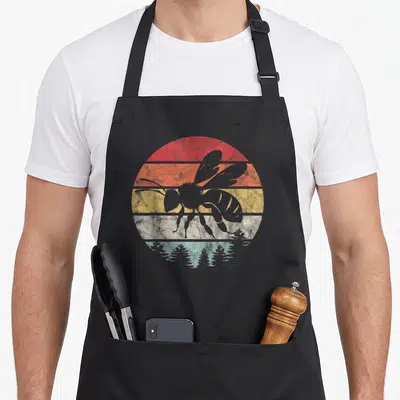 Tablier de cuisine noir avec silhouette d'abeille vintage sur cercle rayé aux couleurs chaudes et silhouettes d'arbres en bas.