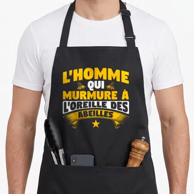 Tablier de cuisine noir avec l'inscription L'homme qui murmure à l'oreille des abeilles en jaune et blanc, agrémenté de deux insectes stylisés et d'une étoile dorée.