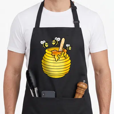 Tablier de cuisine noir avec illustration colorée d'un pot de miel et de trois abeilles souriantes en vol autour.