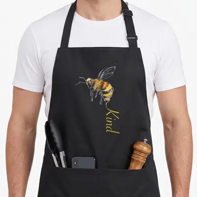 Tablier de cuisine noir avec une illustration d'abeille jaune et noire et le mot Kind écrit en dessous.