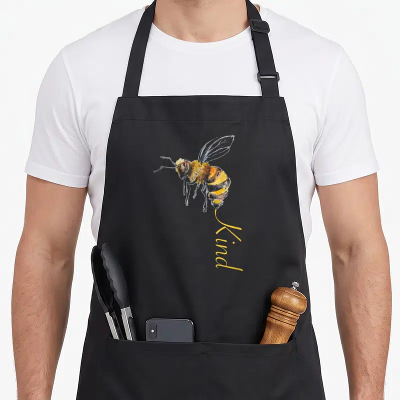 Tablier de cuisine noir avec une illustration d'abeille jaune et noire et le mot Kind écrit en dessous.