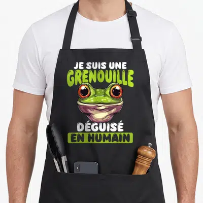 Tablier de cuisine noir avec illustration d'une grenouille verte aux grands yeux orange et texte humoristique sur fond uni.