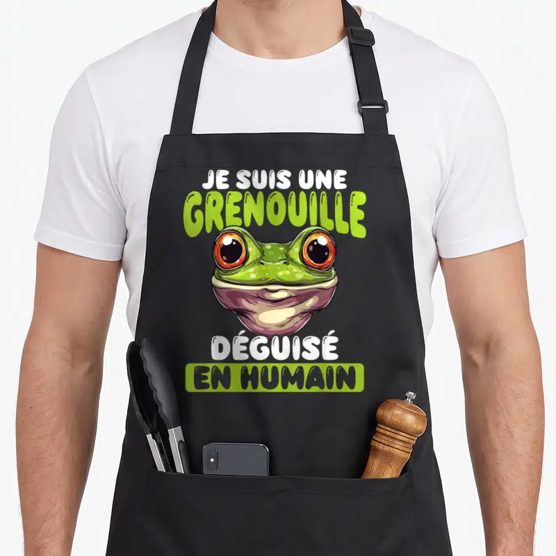 Tablier de cuisine noir avec illustration d'une grenouille verte aux grands yeux orange et texte humoristique sur fond uni.