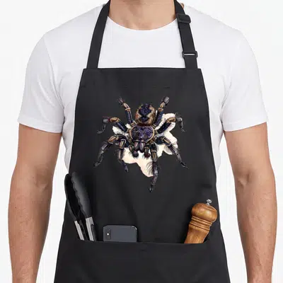 Tablier de cuisine noir avec une grande araignée tarentule aux teintes violettes et brunes sur fond clair.