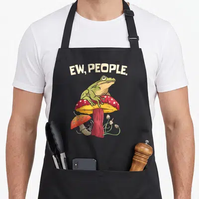 Tablier de cuisine noir avec une grenouille verte assise sur un champignon rouge et l'inscription Ew Gens en haut.