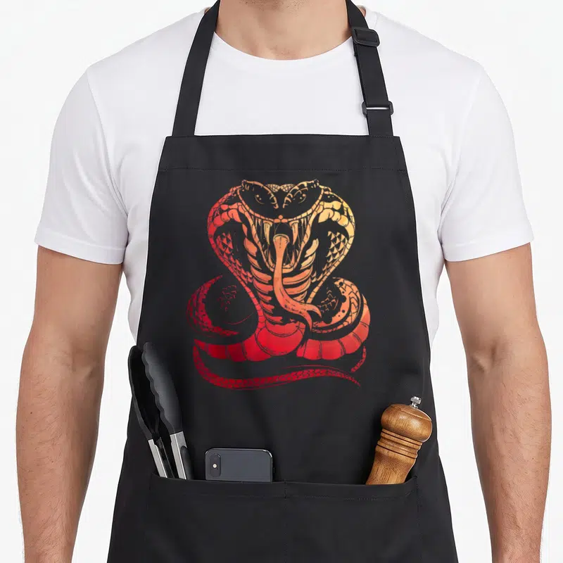 Tablier de cuisine noir avec illustration d'un cobra aux couleurs chaudes rouge et orange, gueule ouverte et langue fourchue visible.
