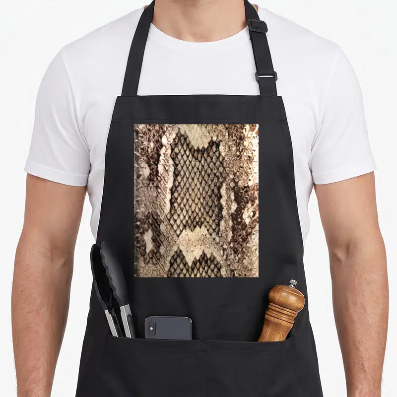 Tablier de cuisine noir avec motif détaillé imitant des écailles de serpent et un motif en losange central beige et brun foncé.